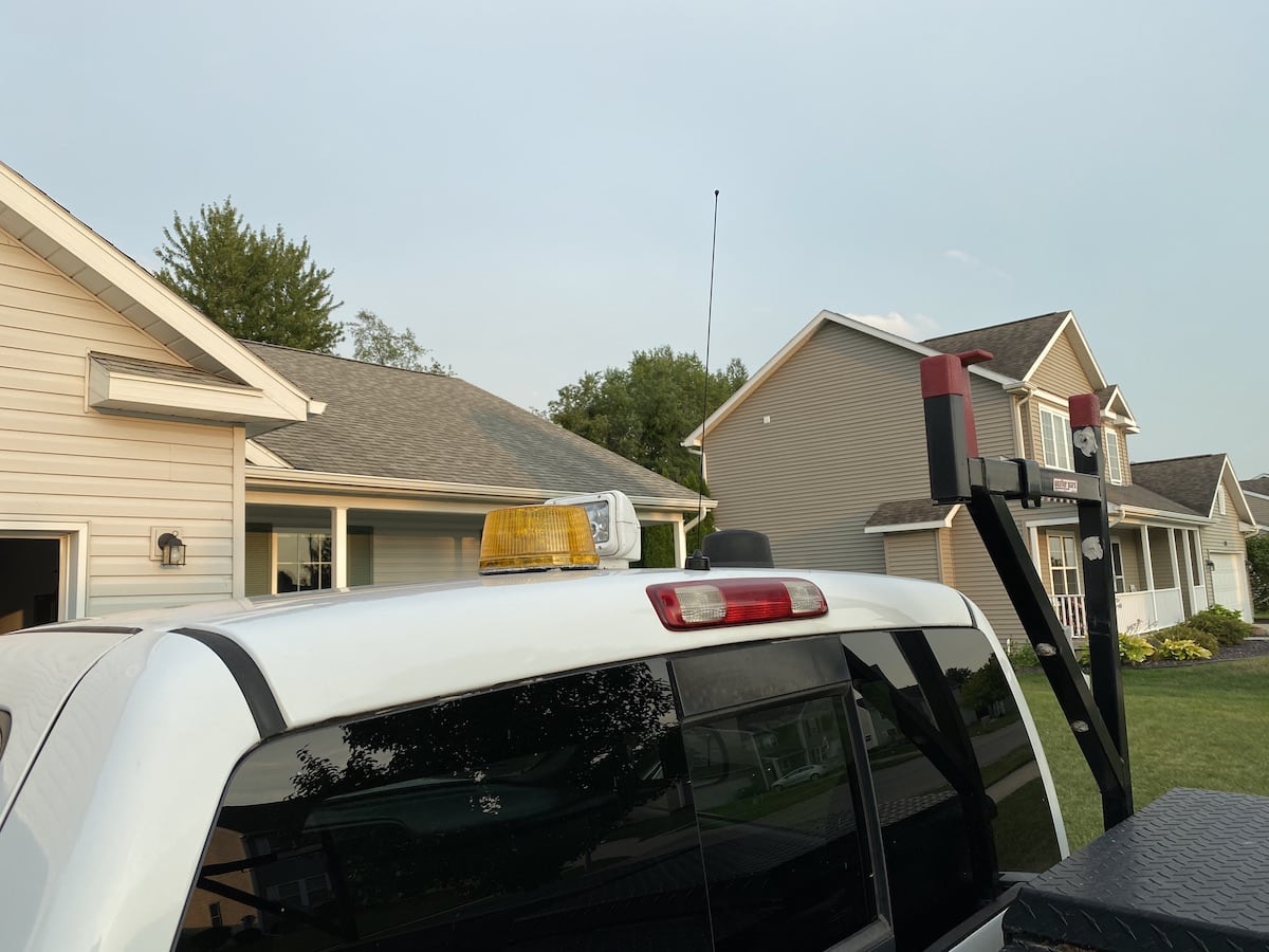 A black
quarter-wave whip antenna flies proudly before a clear summer sky. It is
attached to an NMO mount at the back-center of the roof of a truck cab. Also
visible on the roof are an orange strobe light and a white remote-controlled
spotlight. In the foreground, a little bit of a ladder-rack and toolbox are
visible in the bed of the truck. Further in the background a couple of homes are
visible.