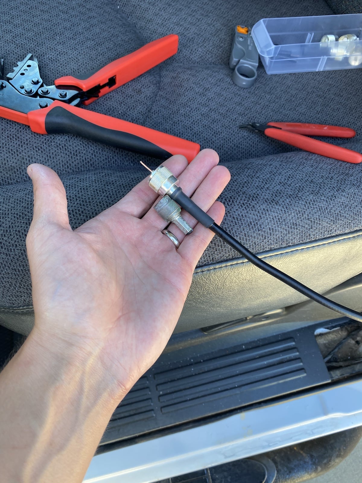 A hand holds
the end of a length of coaxial cable with a UHF connector attached. The center
conductor has not yet been trimmed or soldered. Next to the UHF connector is
an old and corroded TNC connector that has been clipped from the end of its
cable. Visible in the background is a truck seat with some wire cutters, a
crimping tool, and a coaxial cable stripping tool sitting on it.