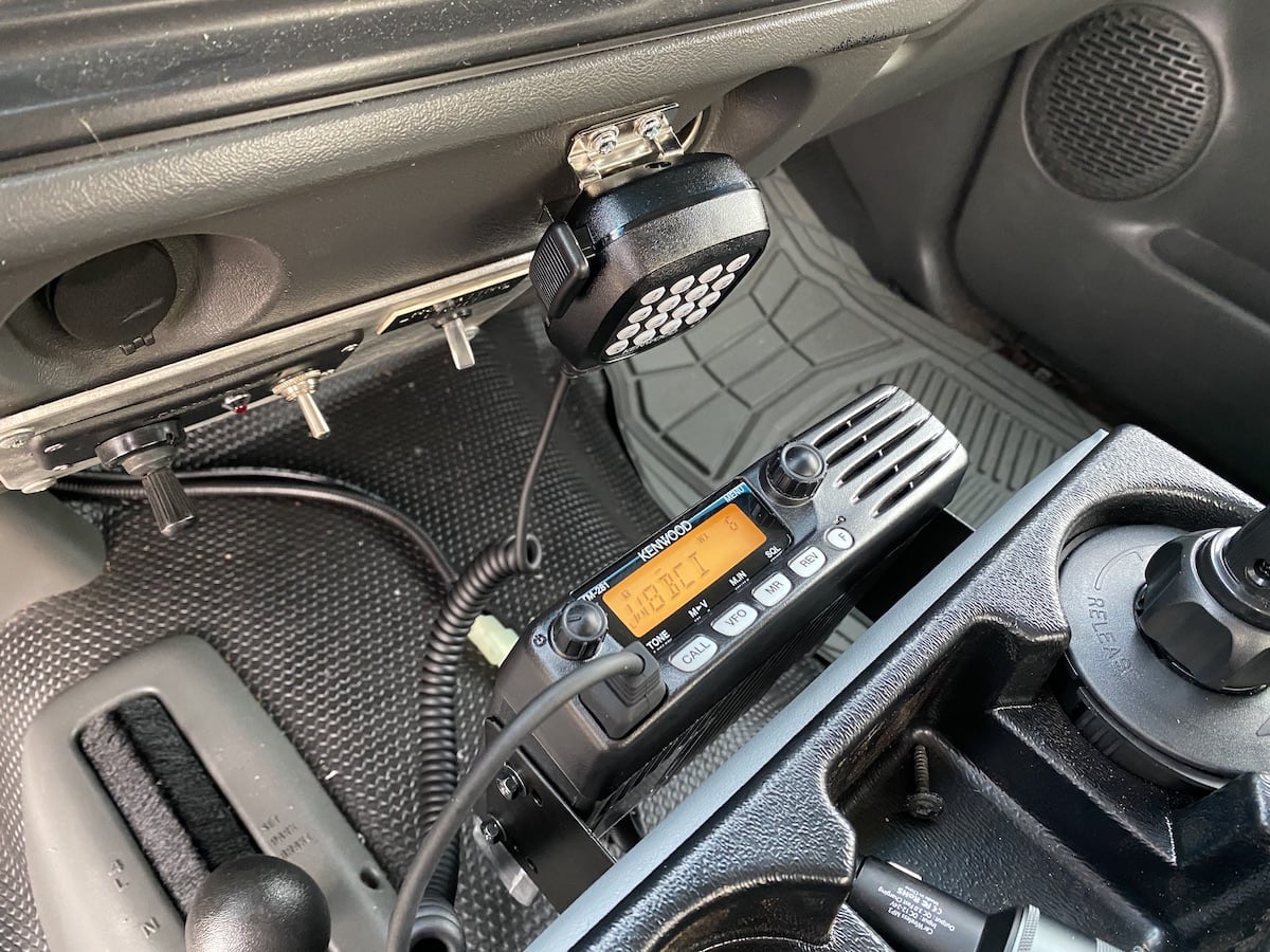 A Kenwood
TM-281a amateur radio transceiver is mounted on the front of an aftermarket
center console in a pickup truck. The radio display is illuminated orange.
In the background, some cables can be seen snaking over the transmission
hump.